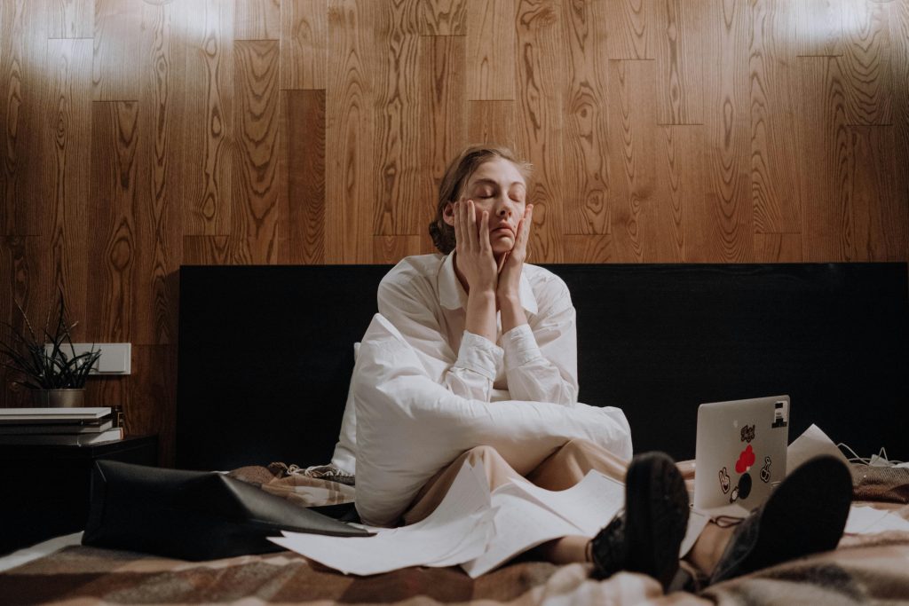 Woman sitting overwhelmed on bed with laptop and papers, indoors.